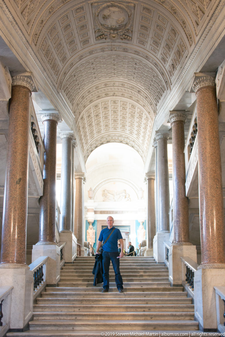 Broad marble staircase with a seated visitor beneath a high coffered barrel vault and rows of reddish granite columns.