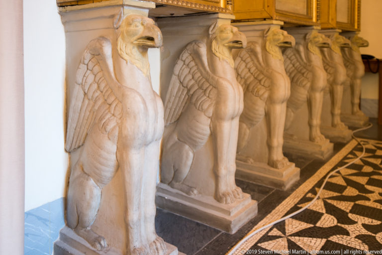 Row of pale carved winged creatures used as supports beneath a stone slab beside a patterned marble floor.