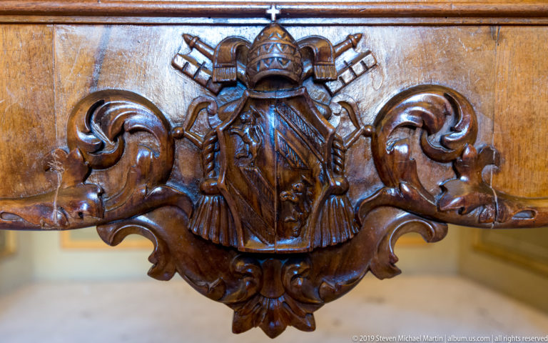Dark carved wood papal emblem with tiered tiara, crossed keys, central shield-like form, and large scrolling ornament.