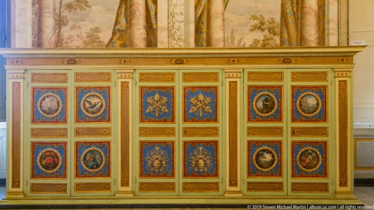 Decorated cabinet with red and blue painted panels, gilded borders, circular medallions, heraldic motifs, and crossed keys.