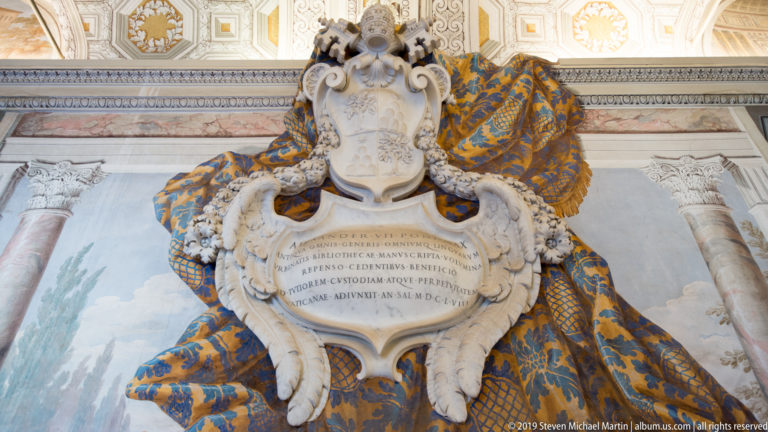 Inscribed marble papal memorial topped by a tiara and heraldic carving, framed by blue-and-gold draped fabric and tassels.