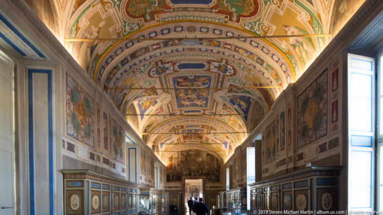 Long Gallery of Maps interior with painted wall maps, ornate cabinetry, tall windows, and a richly frescoed vaulted ceiling.