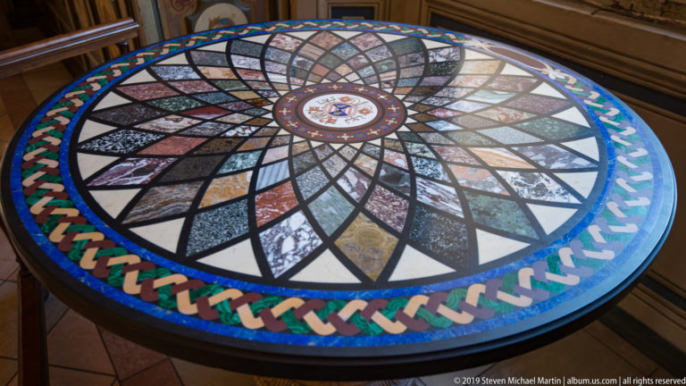 Circular inlaid stone tabletop with radiating multicolored geometric segments, central medallion, black outlines, and braided border.