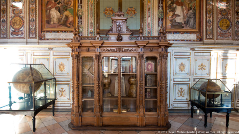 Ornate wooden glass-front cabinet containing instruments, flanked by two large globes in a richly decorated museum room.