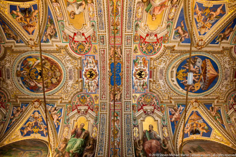 Symmetrical frescoed and gilded ceiling with oval figure scenes, papal shields, seated figures, and dense multicolored ornament.