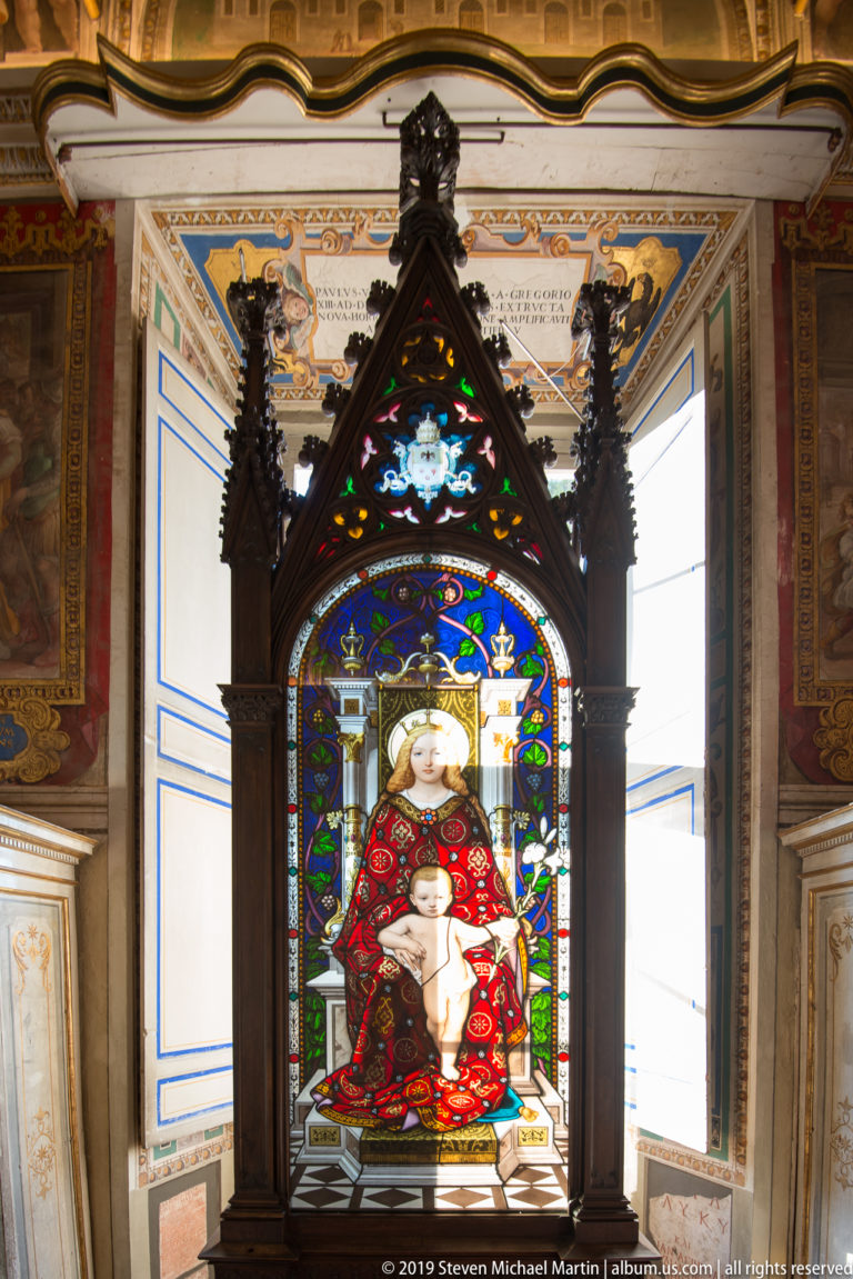 Stained-glass Virgin and Child in vivid red, blue, green, and gold within a dark pointed Gothic-style frame.