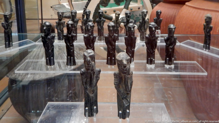 Rows of slender dark Egyptian statuettes in a glass case, with two larger standing figures positioned at the front.