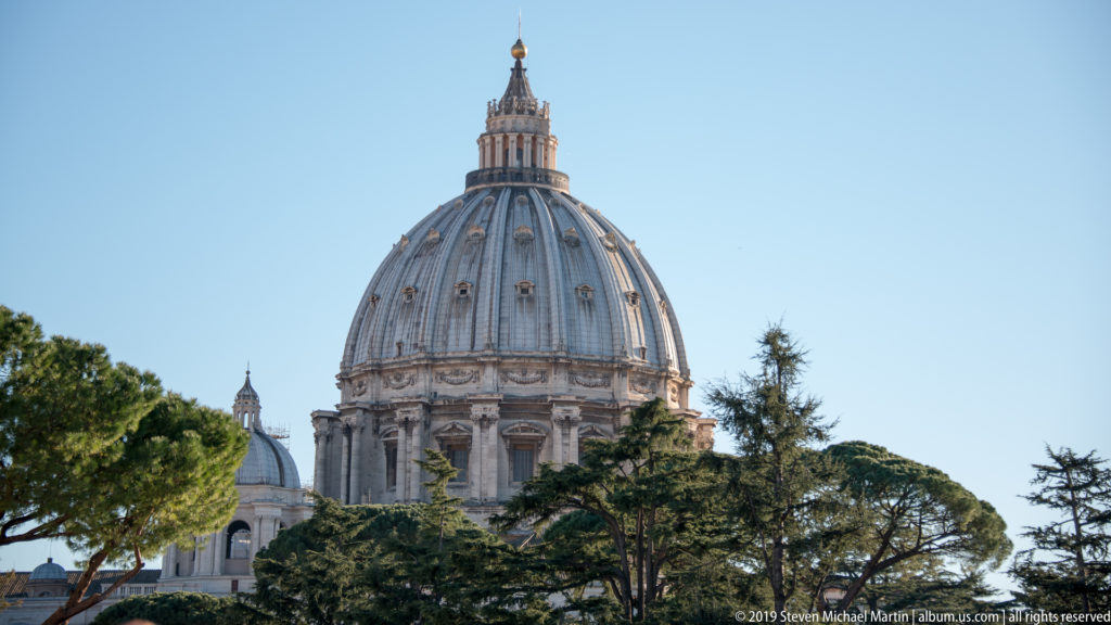 St. Peter's Basilica dome rising above mature trees under a clear blue sky, with its lantern and gilded finial visible.