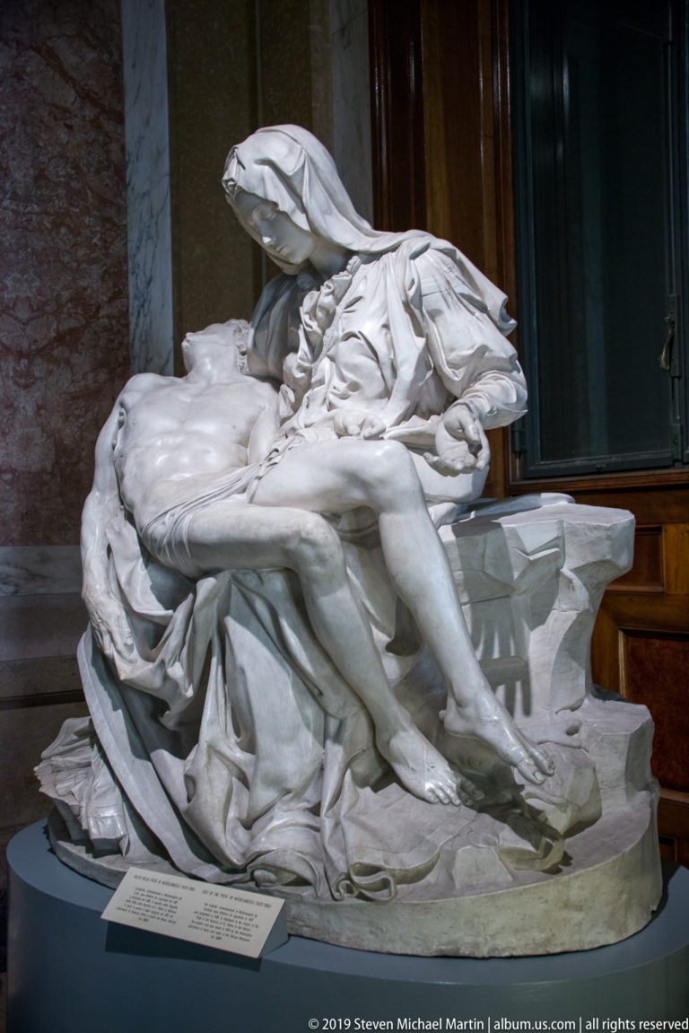 Side view of a white marble Pietà with Christ's body stretched across the lap of the veiled Virgin.