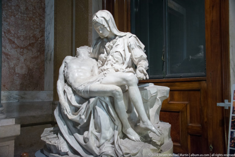 Three-quarter view of a white marble Pietà showing the veiled Virgin supporting Christ's limp body across her lap.