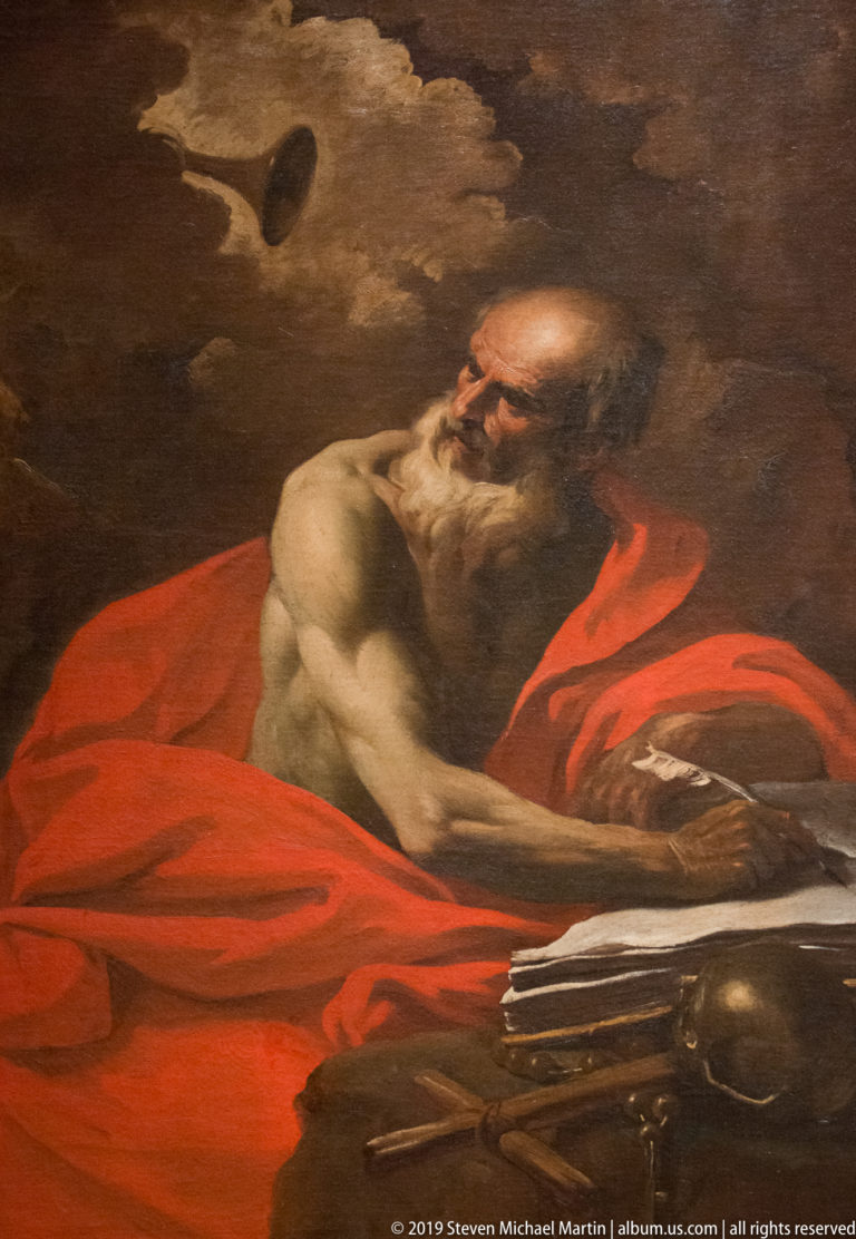 Elderly bearded saint in a red mantle leans over an open book and writing surface against a dark background.