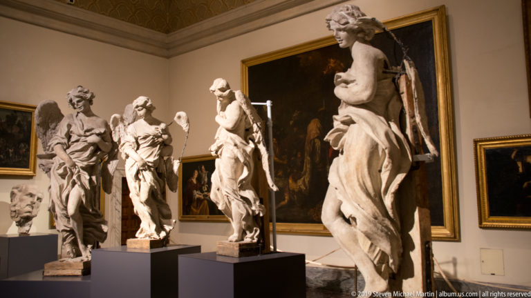 Gallery with several life-size marble statues on dark pedestals arranged in front of large framed paintings.