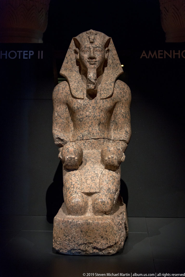 Seated pink-granite Egyptian pharaoh wearing a broad striped headdress, hands resting on thighs against a dark background.