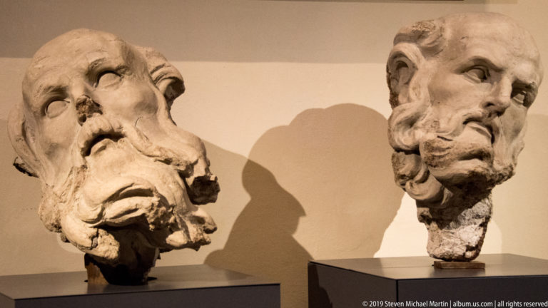 Two ancient marble male heads on separate mounts, one heavily damaged and one more complete with a curling beard.