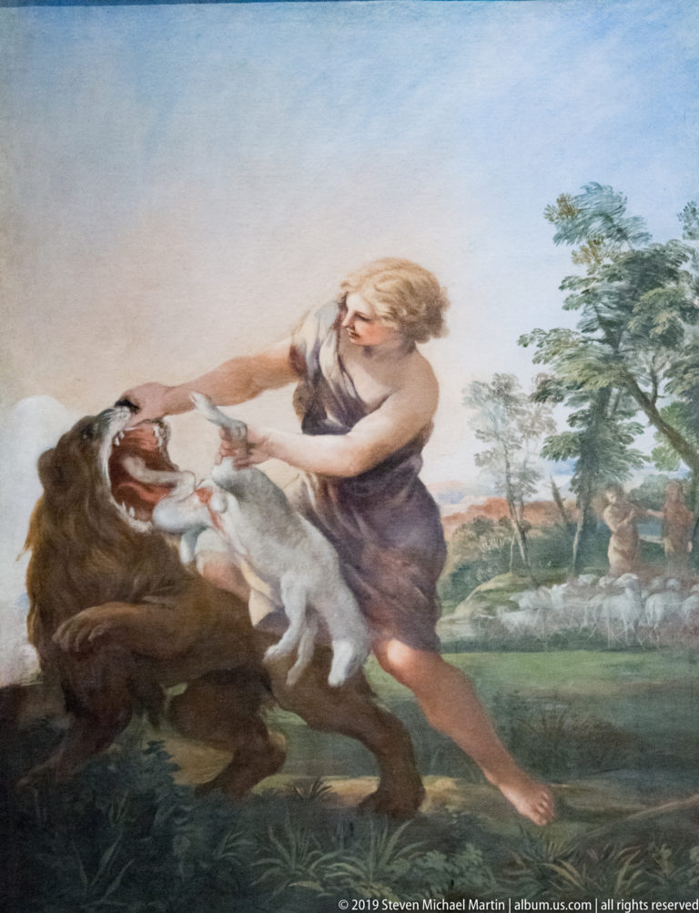 Painted scene of a muscular man wrestling a lion in a pastoral landscape with trees and grazing animals behind.