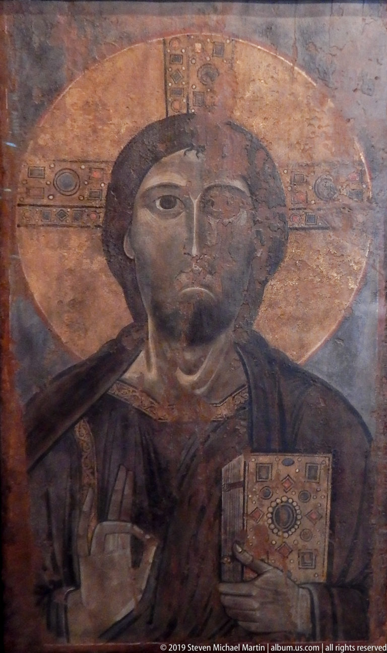 Frontal medieval image of Christ with a cruciform halo, right hand raised in blessing, and jeweled book held to his chest.