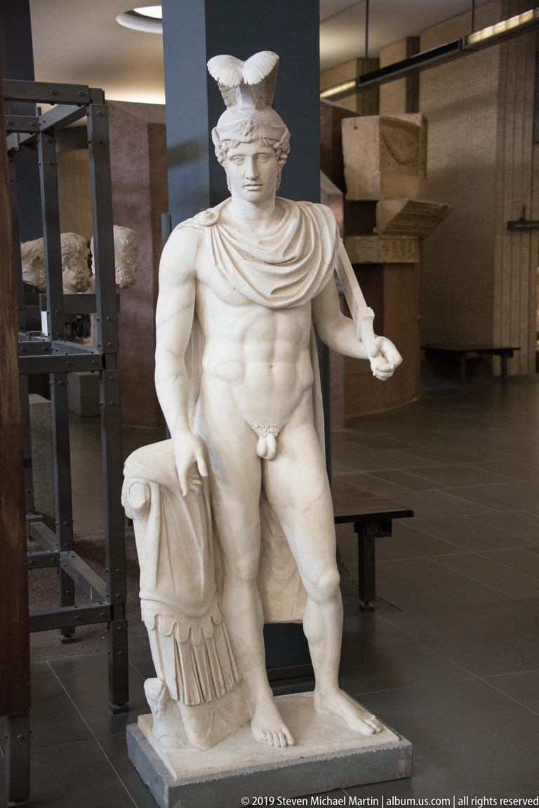 Marble statue of a nude male figure wearing a high crested helmet and drapery, with one arm extended outward.