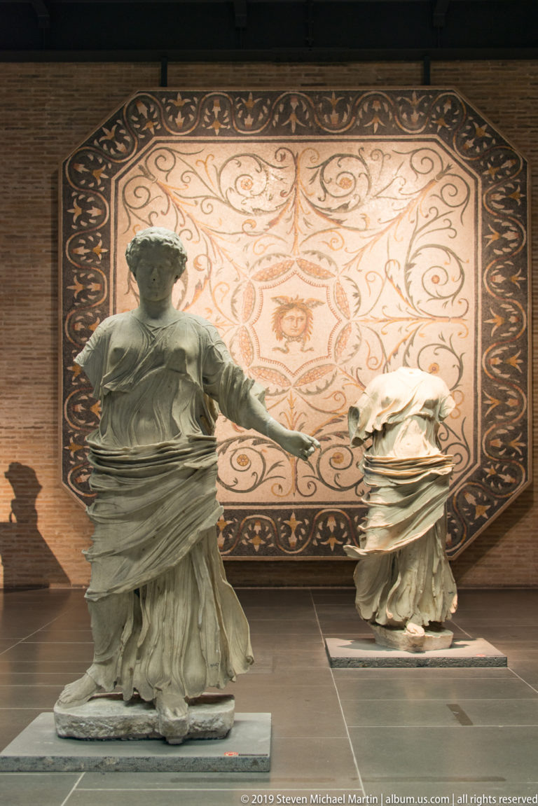Two headless draped marble statues stand before a large wall-mounted mosaic with scrolling vine ornament and a central motif.