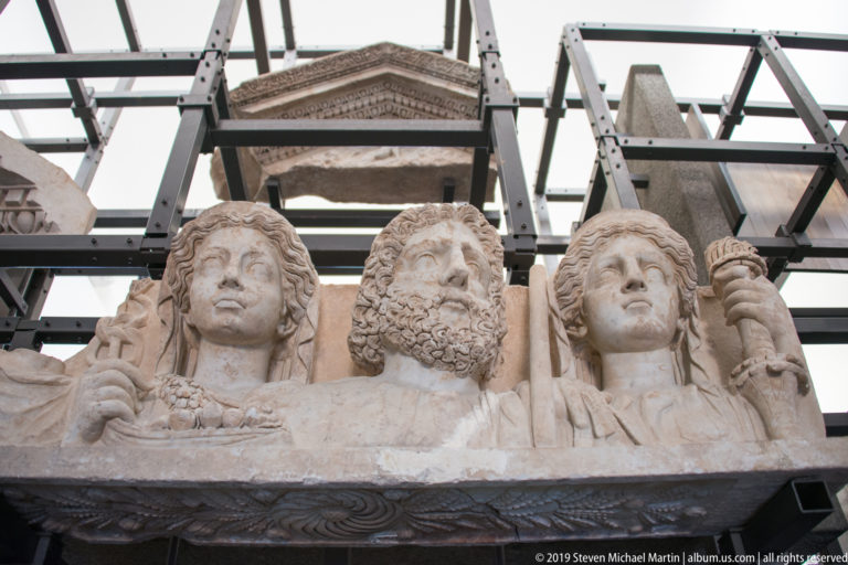 Three carved marble bust-length figures, including a central bearded man, project above an ancient stone monument.