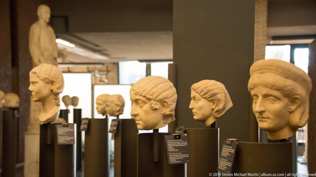 Row of ancient marble portrait heads on dark stands, with varied hairstyles and facial features receding through a museum gallery.