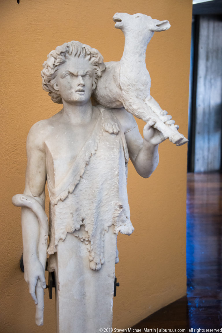 Fragmentary marble statue of a youthful figure wearing an animal skin and carrying a small hoofed animal over one shoulder.