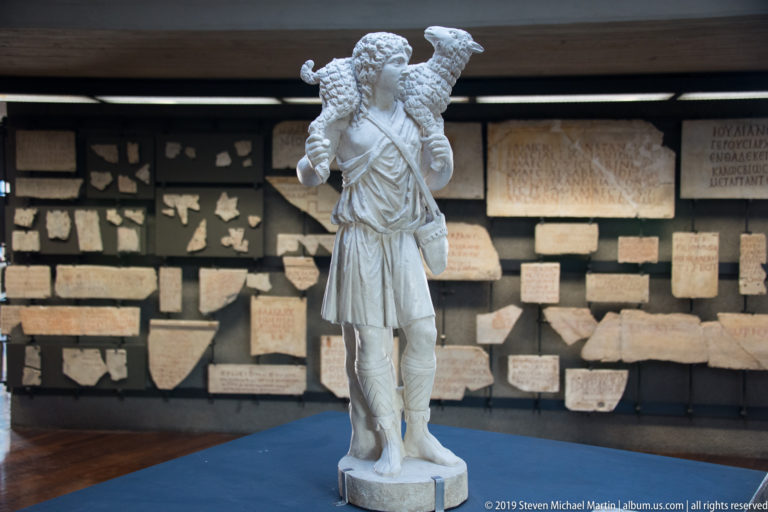Marble statue of a youthful shepherd in a short tunic carrying a lamb across his shoulders before a wall of inscriptions.