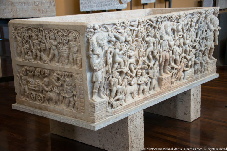 Large marble sarcophagus densely carved with overlapping human figures, animals, weapons, and drapery across its side and end.