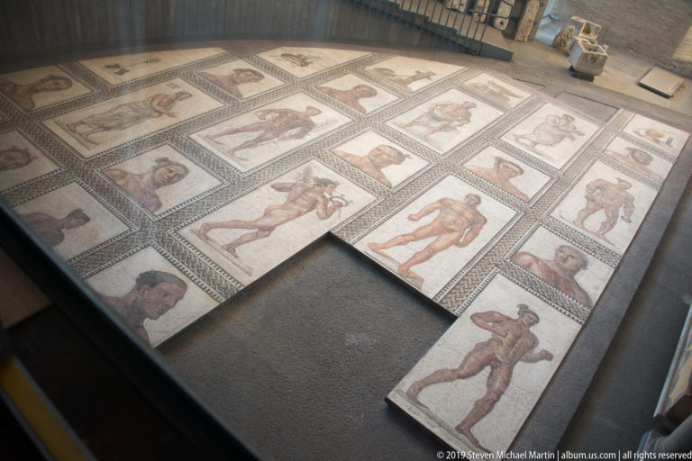 Large reconstructed mosaic pavement with athletes, heads, geometric borders, and several missing rectangular sections.