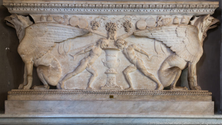 Pale marble relief with two symmetrical winged animals facing a tall central pedestal, with fruit-like ornament above.