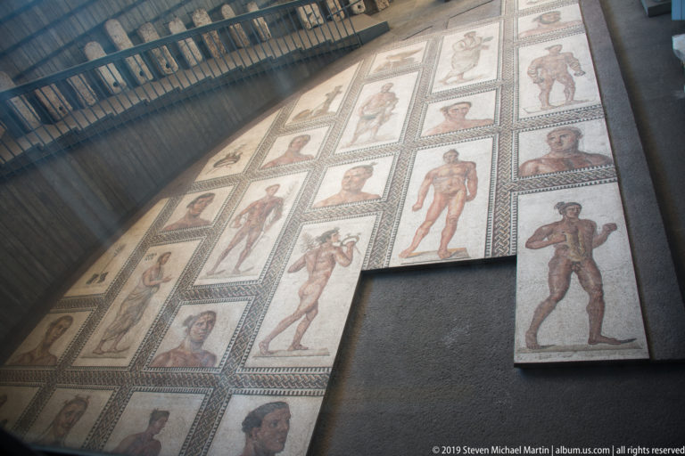 Oblique view of a large mosaic pavement with rectangular panels of athletes and portrait heads separated by geometric borders.
