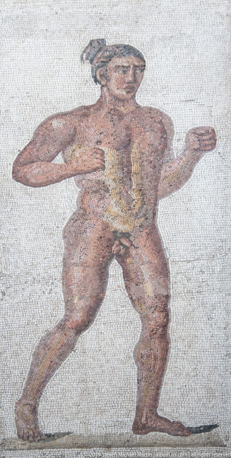 Ancient mosaic of a muscular nude male figure striding with bent arms across a pale tessellated background.