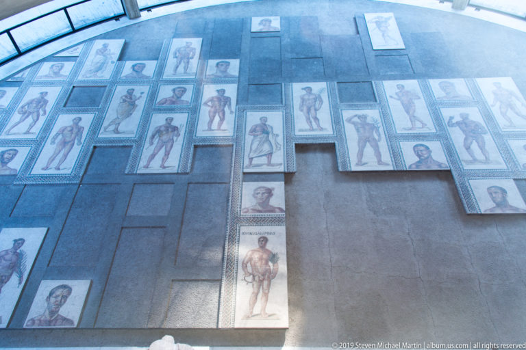 Overhead view of many rectangular ancient mosaic panels showing standing human figures and portrait heads on a dark display surface.