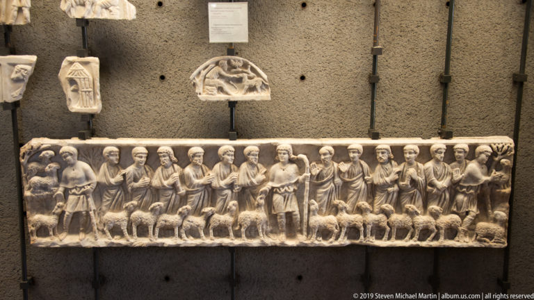 Long marble relief with a row of standing figures and sheep, displayed beneath smaller carved fragments on a dark wall.