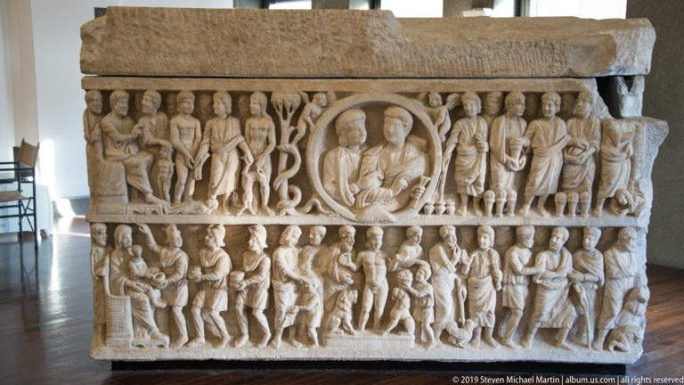 Marble sarcophagus carved in two registers with crowded standing figures, narrative scenes, and a central framed figure.