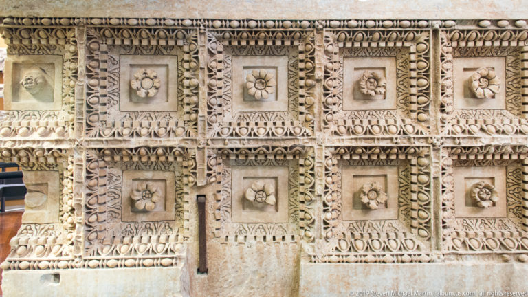 Carved pale stone panel divided into square fields with rosettes, masks, inscriptions, beadwork, and repeating geometric ornament.