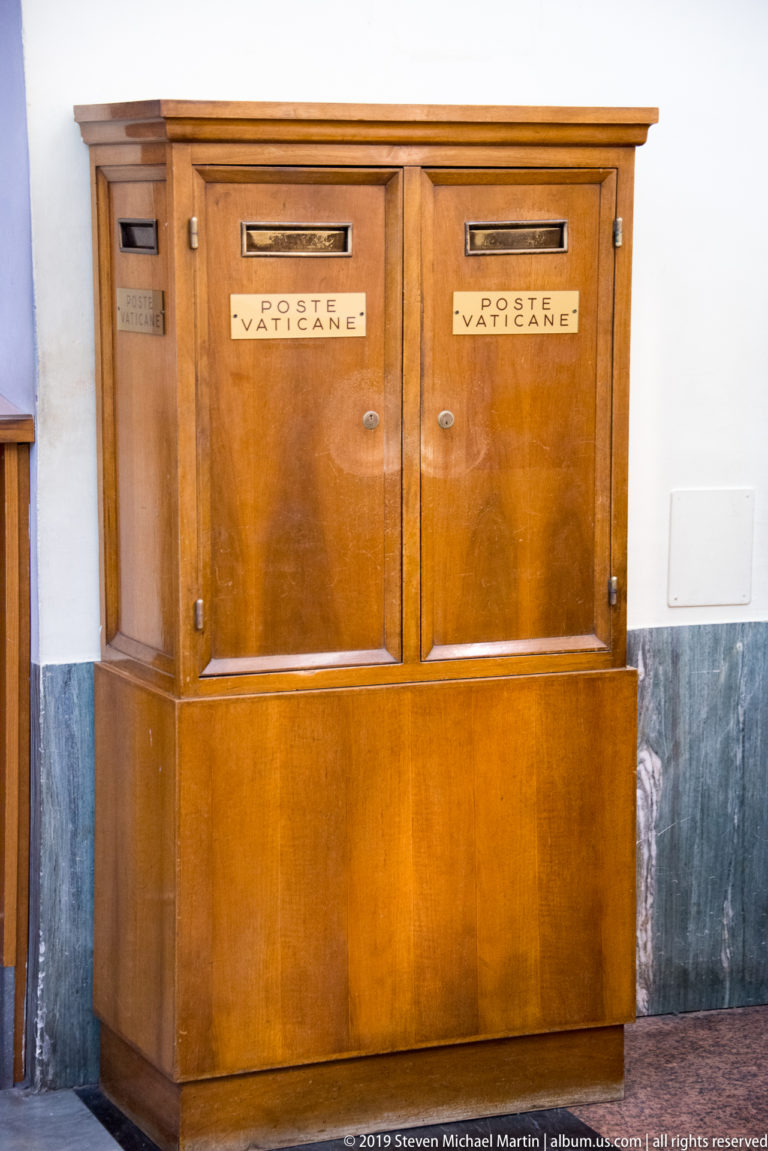 Wooden Vatican postal cabinet with two openings labeled POSTE VATICANE against a pale wall and dark marble dado.