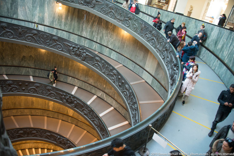 Visitors descend the curved double-helix ramps of Giuseppe Momo's Vatican Museums staircase beside dark relief-patterned balustrades.