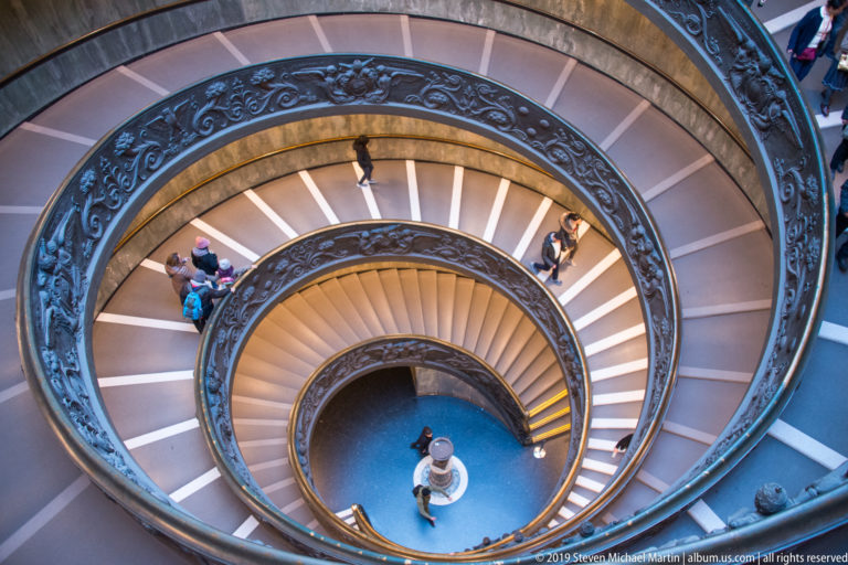 Top-down view of Giuseppe Momo's Vatican Museums staircase with visitors on concentric double-helix ramps surrounding the central well.