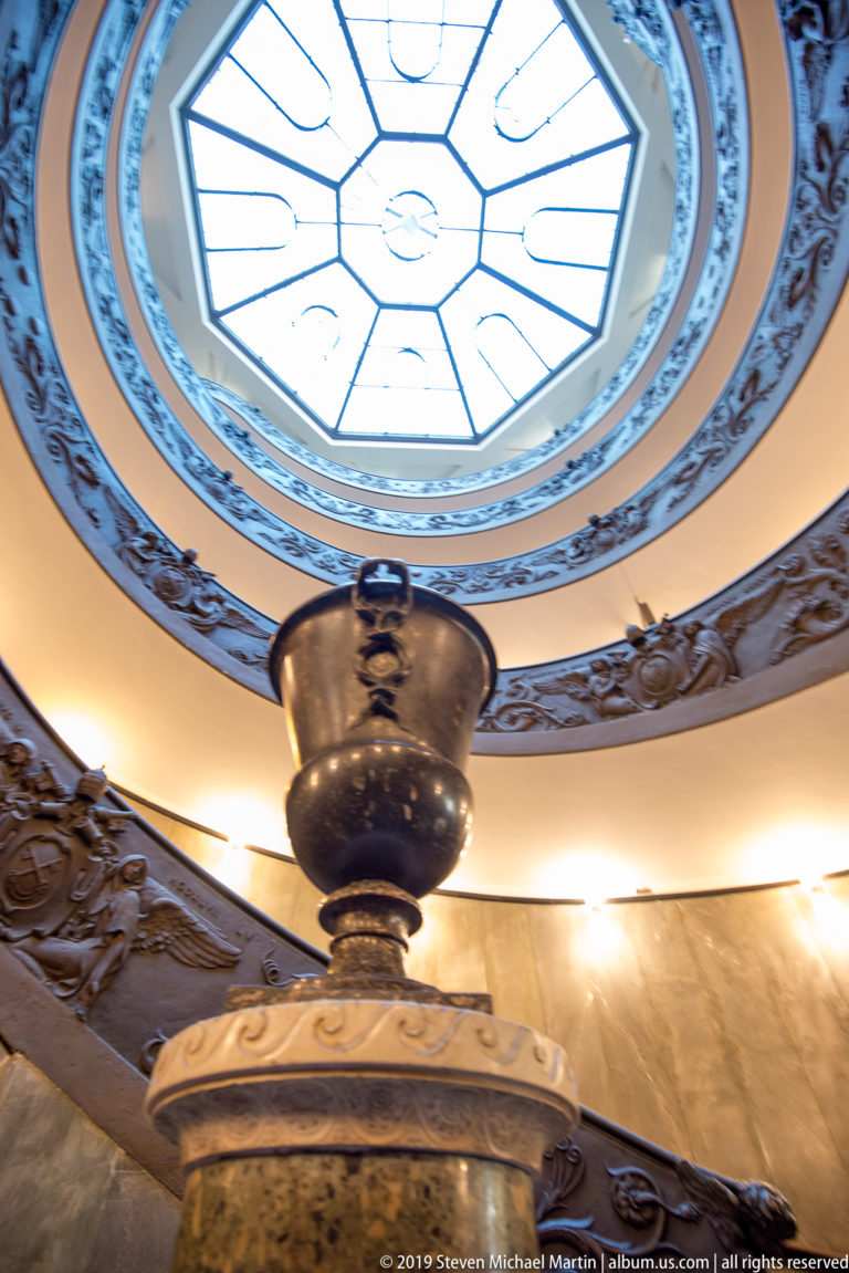 Large decorative urn beneath the curving double-helix ramps and bright geometric skylight of Giuseppe Momo's Vatican Museums staircase.