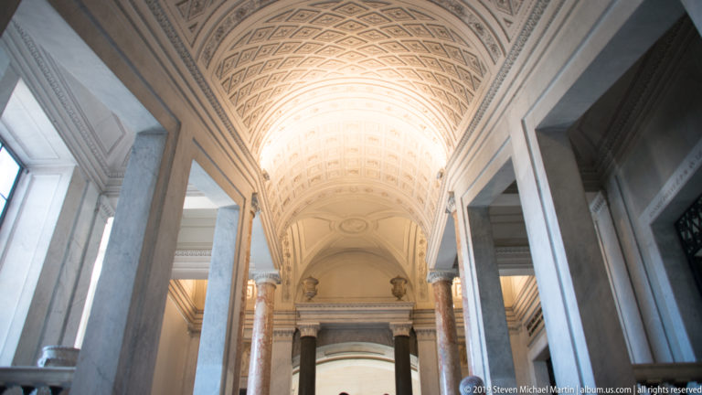 Long pale museum corridor with coffered barrel vault, marble columns and pilasters, side openings, and a bright distant doorway.