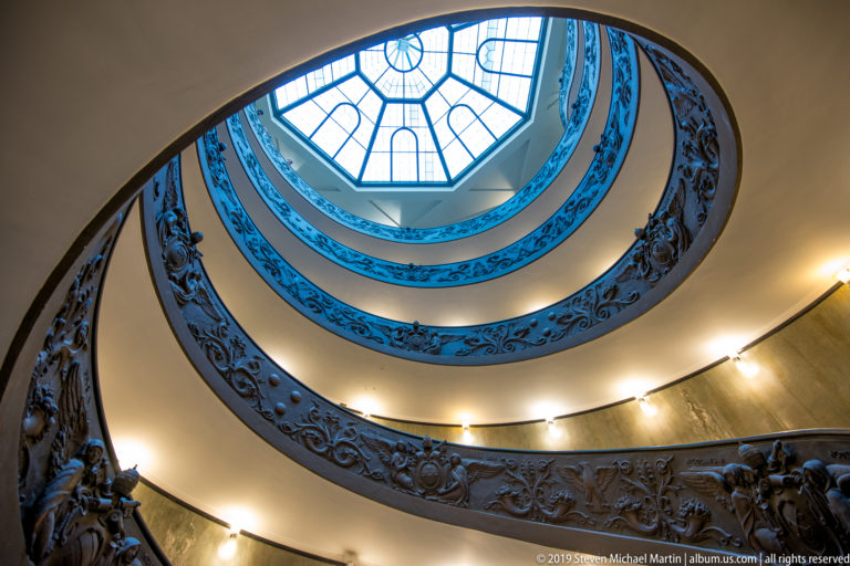 Concentric ramps of Giuseppe Momo's Vatican Museums staircase curve beneath a geometric skylight, edged by dark ornamental balustrades.