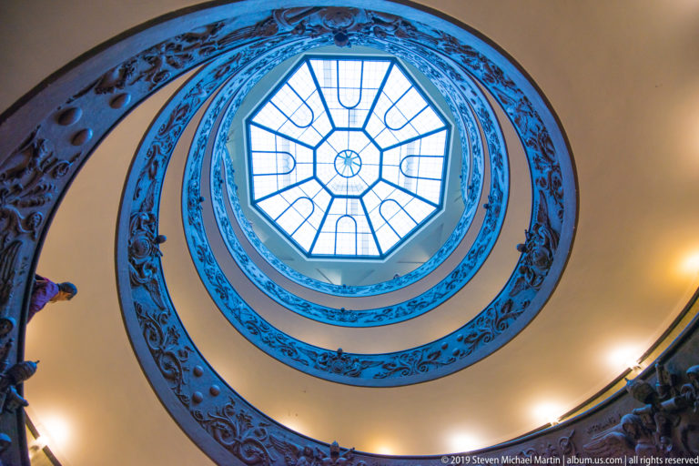 Upward view through Giuseppe Momo's double-helix staircase in the Vatican Museums, with ornate balustrades curving toward a geometric skylight.