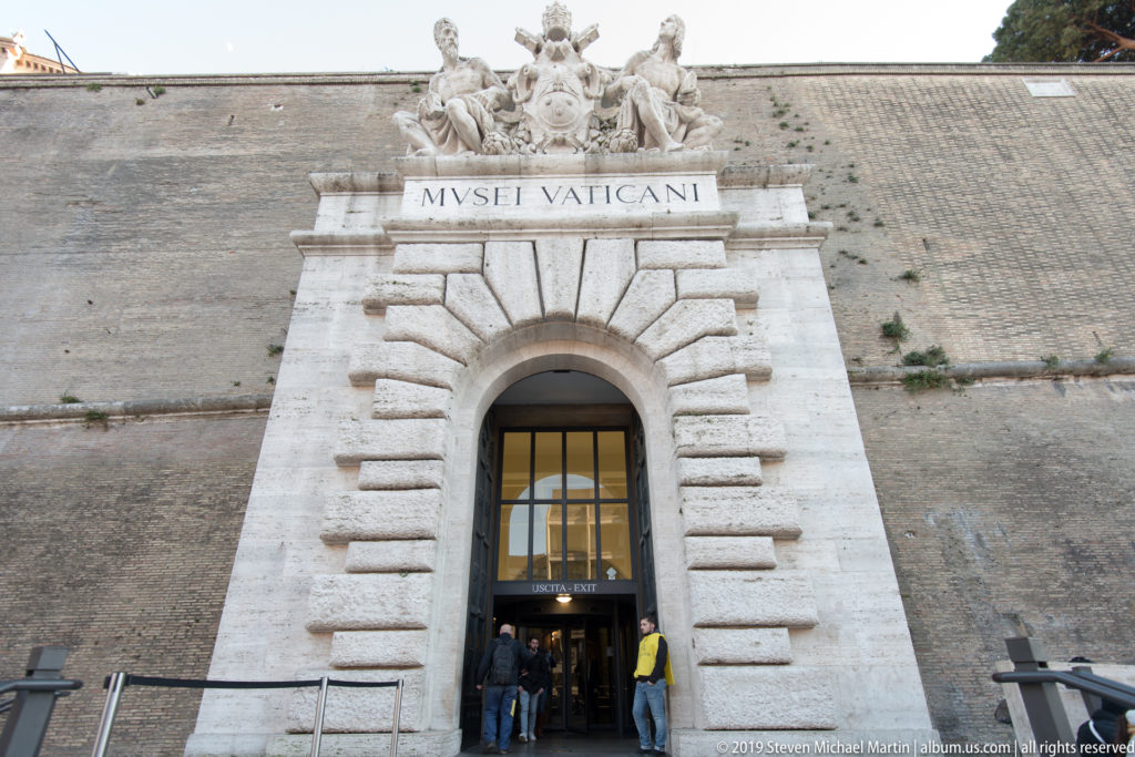 Vatican Museums entrance with rusticated stone arch, MVSEI VATICANI inscription, crowning sculpture, and visitors at the doorway.