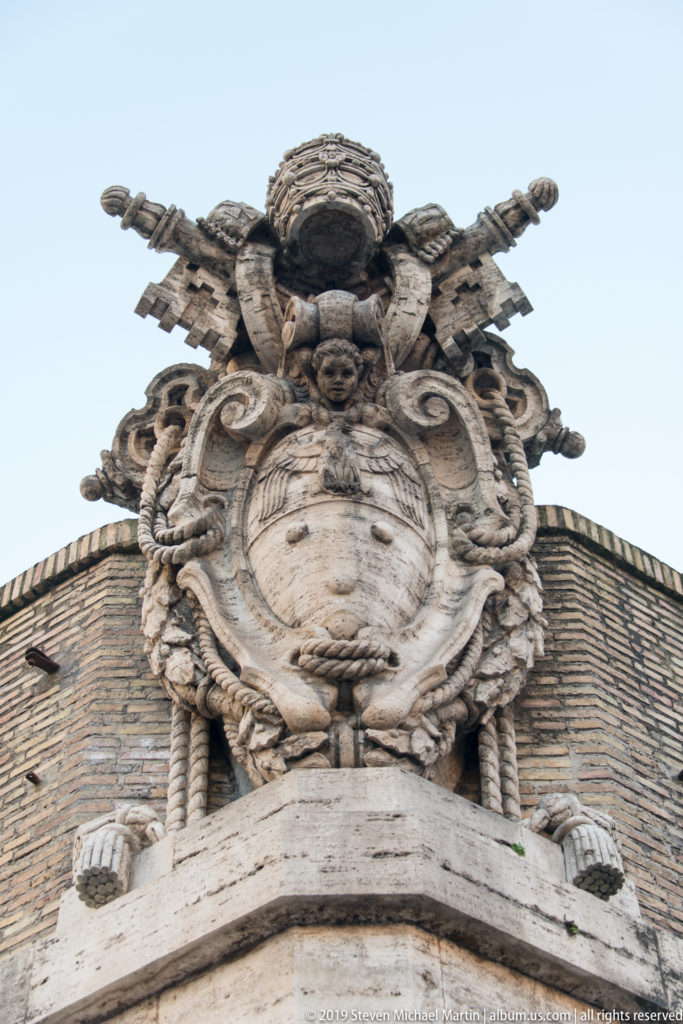 Large carved papal coat of arms with shield, scrollwork, crossed keys, and tiara above a brick Vatican Museums wall.