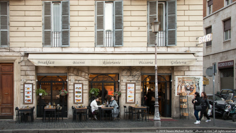Restaurant storefront with outdoor tables, menu boards, pedestrians, shuttered windows, and parked vehicles on a street near the Vatican Museums.