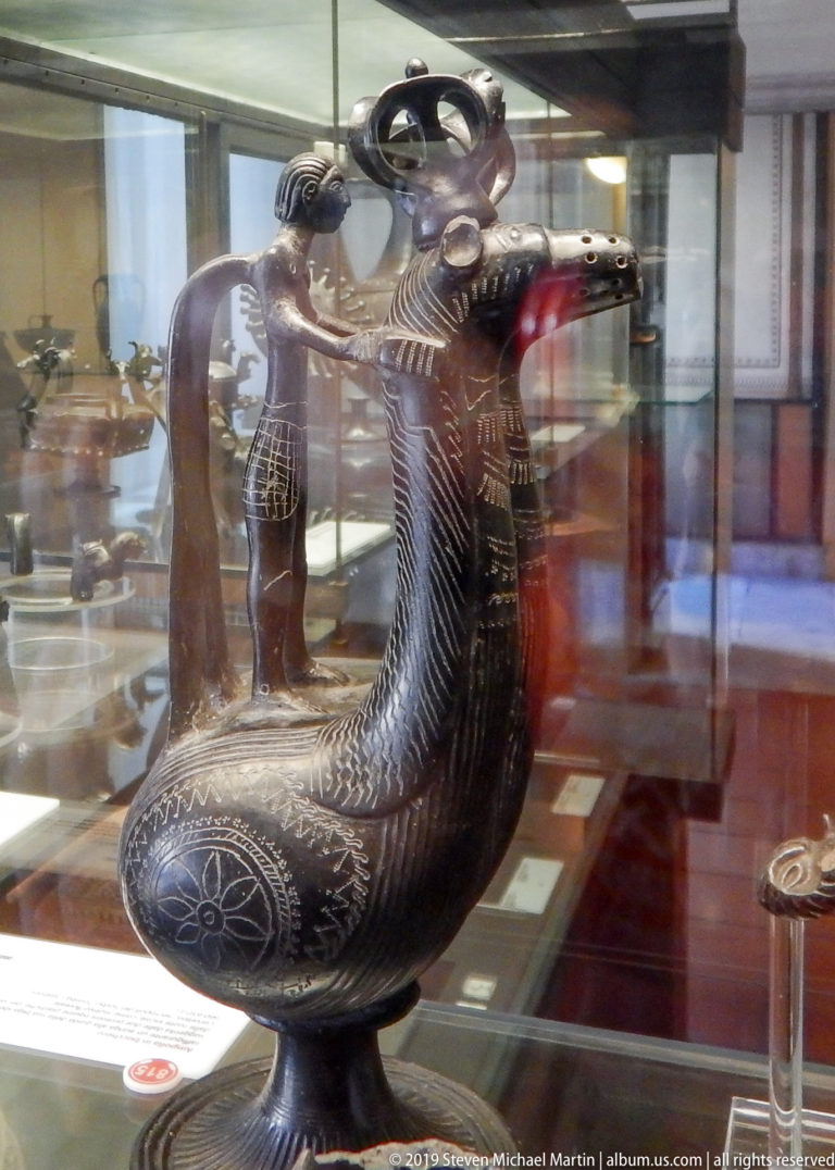 Dark metal bird-shaped vessel with open beak spout, incised feather patterns, rounded body, and tall looping handle.