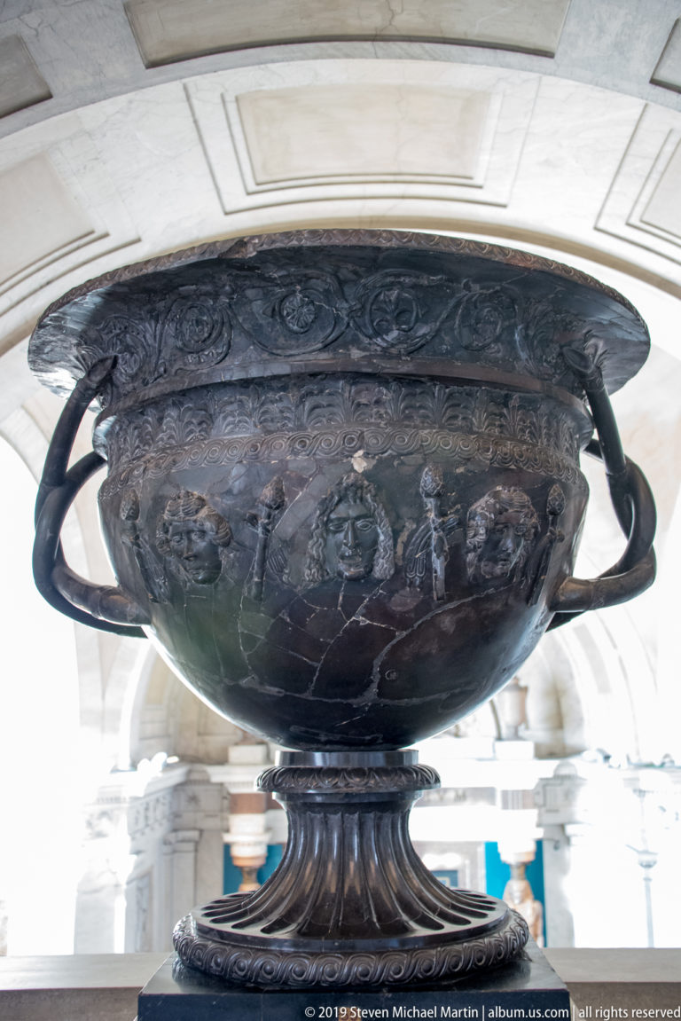 Large dark metal krater with broad rim, loop handles, fluted foot, and figural relief decoration across its rounded body.