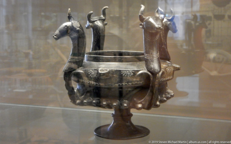Dark metal bowl on a short foot with several horned animal heads projecting around the rim and incised exterior decoration.