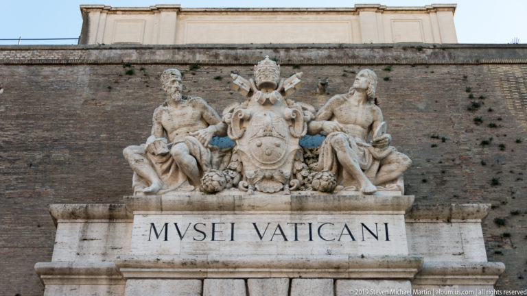 Stone Vatican Museums crest with papal tiara, crossed keys, central shield, two reclining male figures, and MUSEI VATICANI inscription.