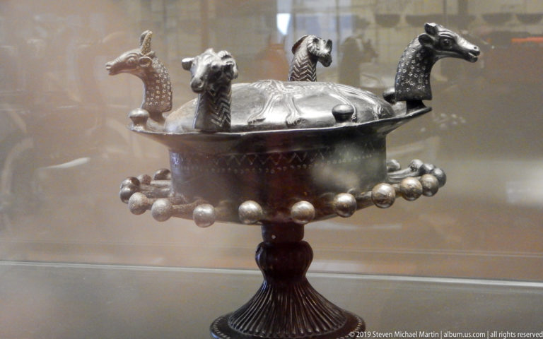 Dark metal pedestal bowl with projecting animal heads, rounded bosses, incised decoration, and a tall fluted foot.
